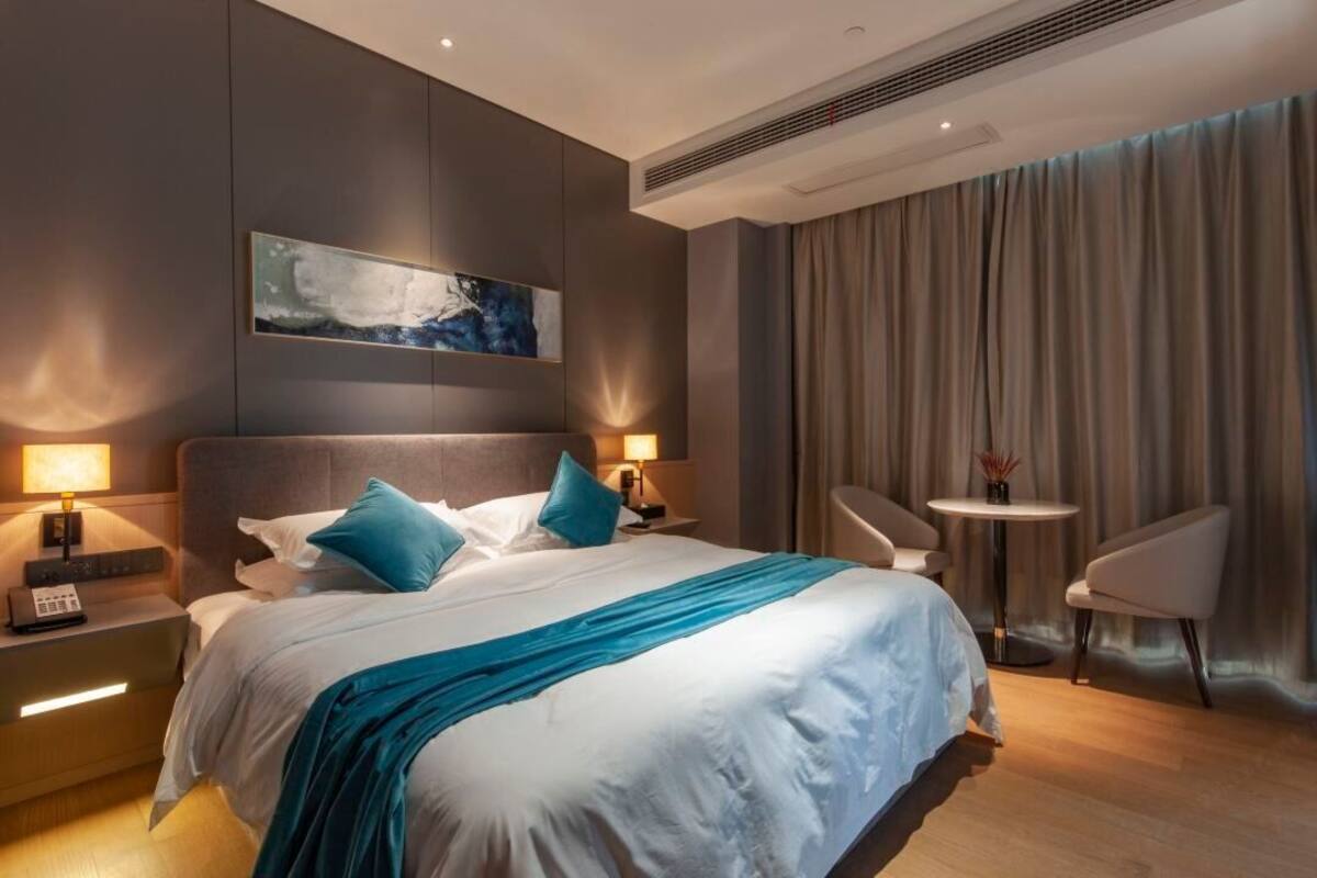 A modern hotel room with a large bed, two bedside tables with lamps, a small round table with a plant, and a chair.
