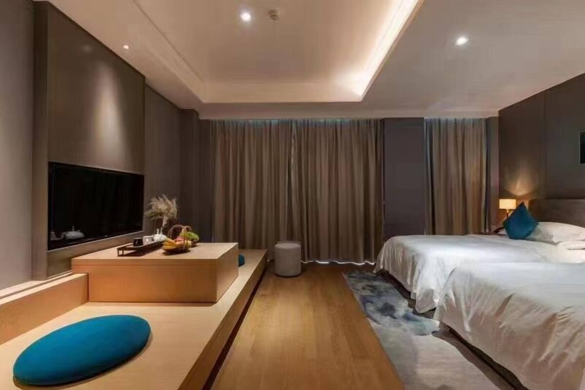 A modern hotel room with a large bed, a TV, a small seating area, and a large window with curtains.