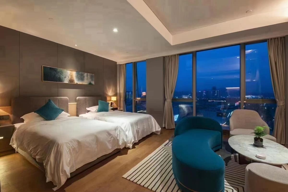 A modern hotel room with two beds, a sitting area with a round table, and a large window offering a city view.