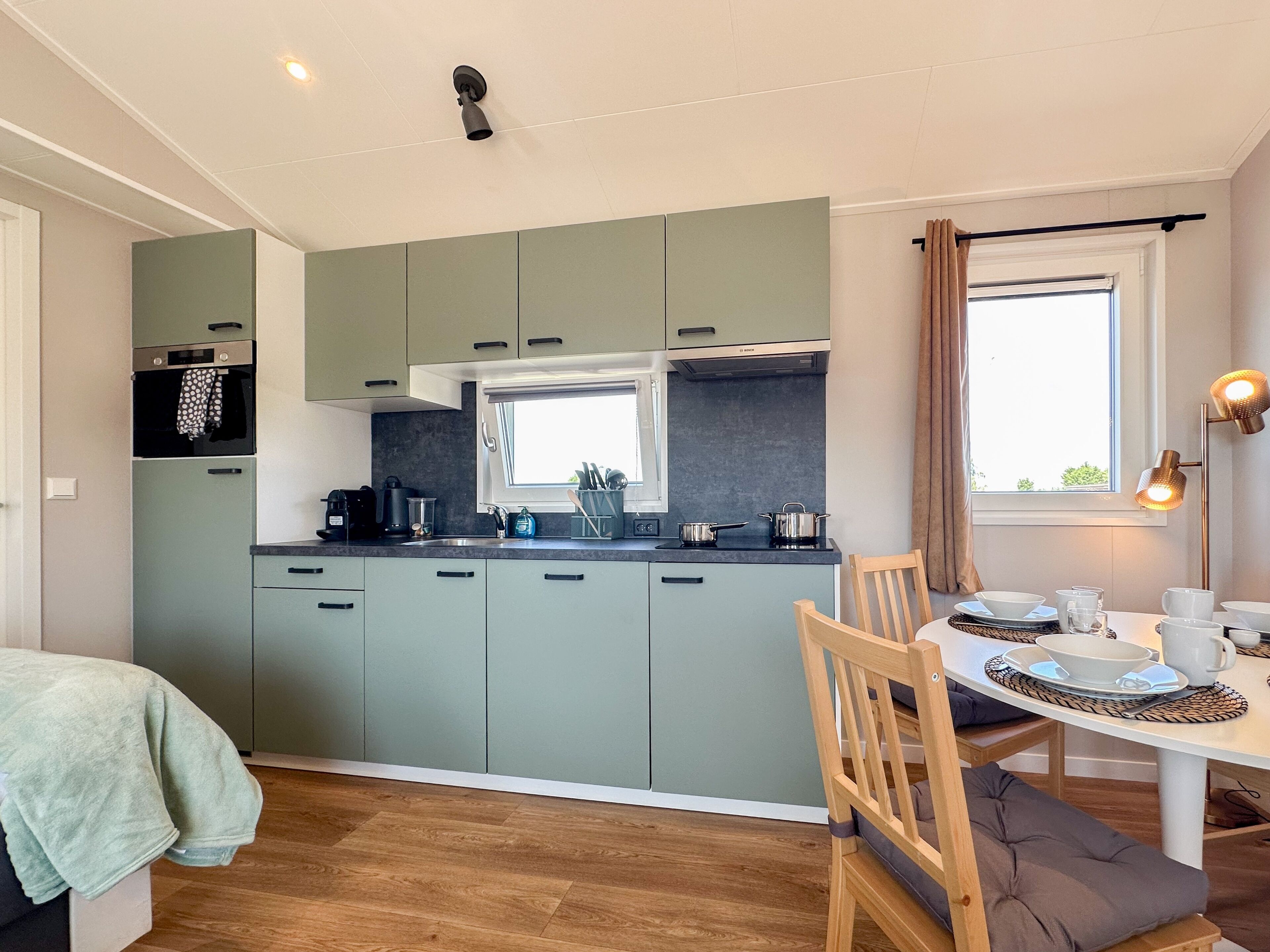 A compact kitchen with a dining area, featuring a window with curtains, a table set with cups and bowls, and a ceiling light.