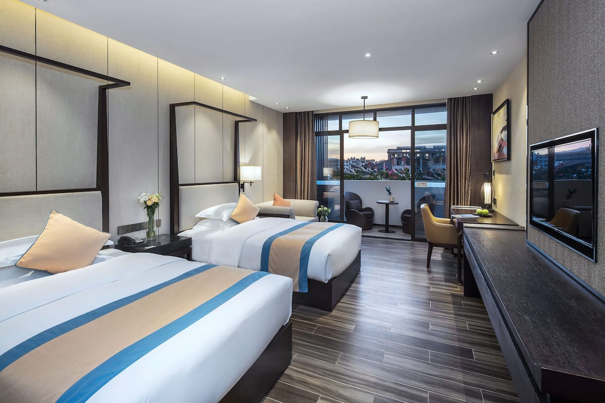 Ein Hotelzimmer mit zwei Betten, einem Schreibtisch, einem Sessel und einem Fernseher, mit Blick auf die Stadt.