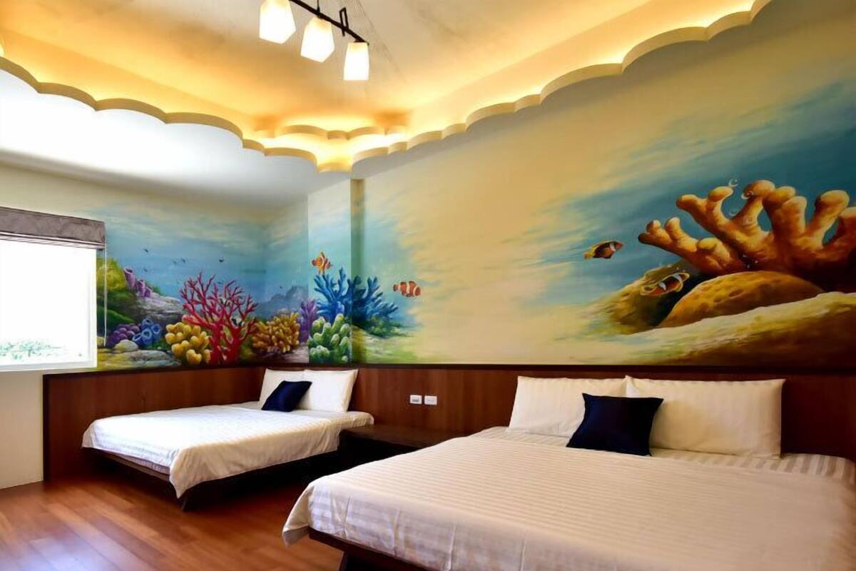 Una habitación de hotel con dos camas, un mural de una escena submarina en la pared y una ventana con vista al mar.