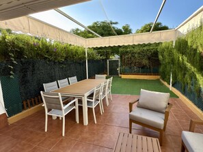 Un patio techado con una mesa y sillas de madera, rodeado por un seto verde y una valla de madera.