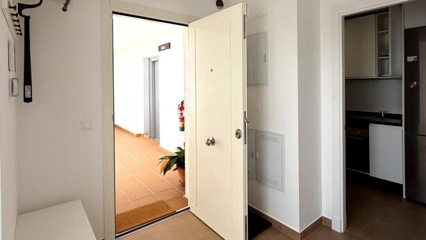 Eine geöffnete Tür führt in einen Flur mit Fliesenboden, einem Blumentopf und einem sichtbaren Feuerlöscher.