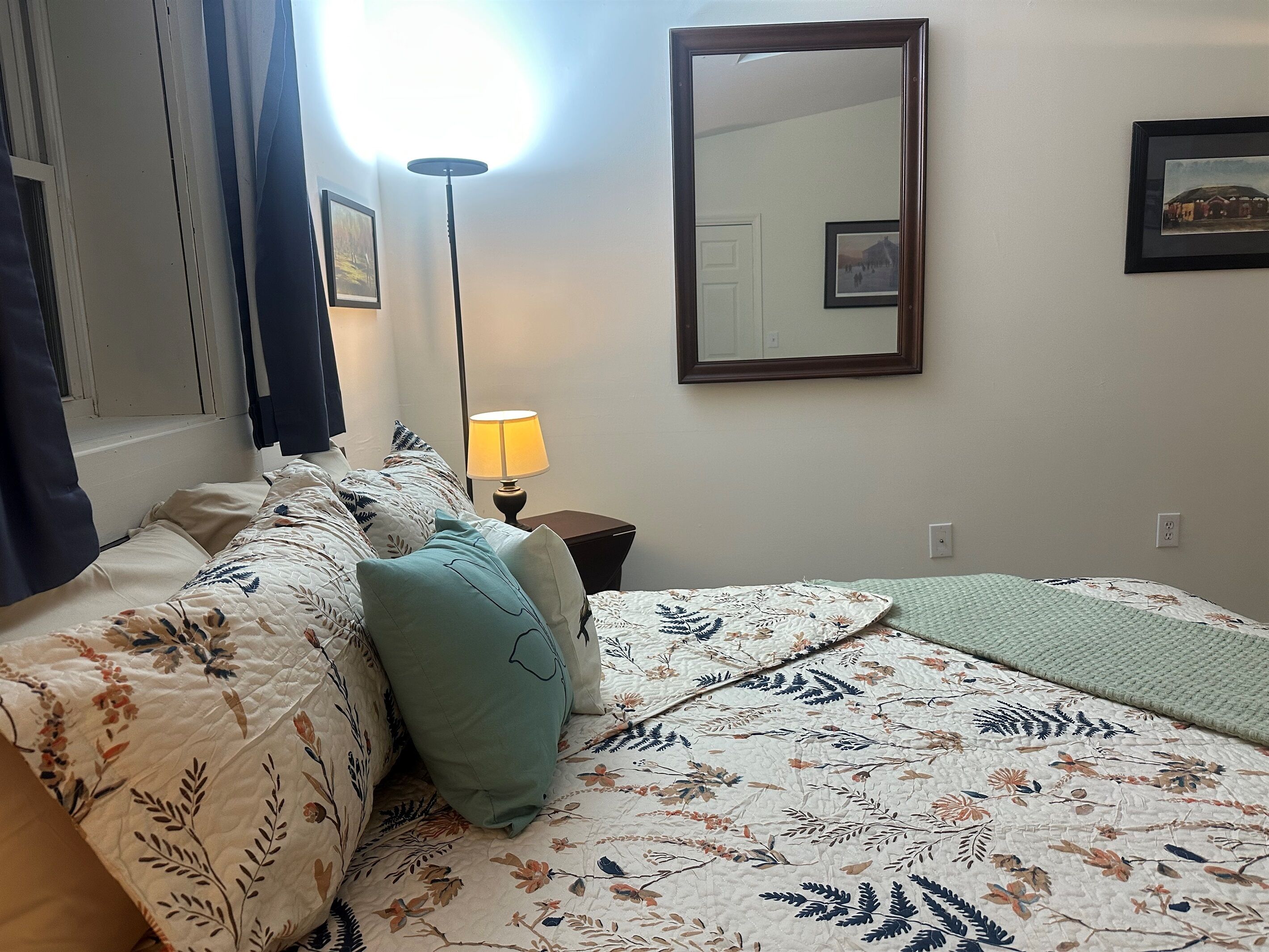Une chambre d’hôtel avec un lit, une table de chevet, une lampe, un miroir et un tableau encadré au mur.
