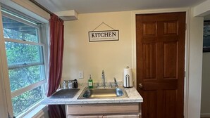 A compact kitchen with a sink, countertop, and wooden cabinets. A 'KITCHEN' sign is on the wall, and a wooden door is visible. - Meadowlark Suite at Cooperstown Lodge 3Miles toCDP (Cooperstown)