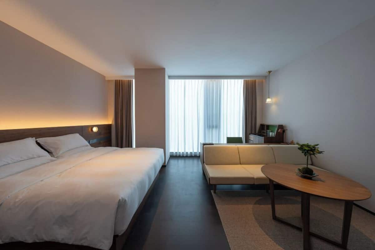 A modern hotel room with a large bed, a sofa, a small table, and a window with curtains.