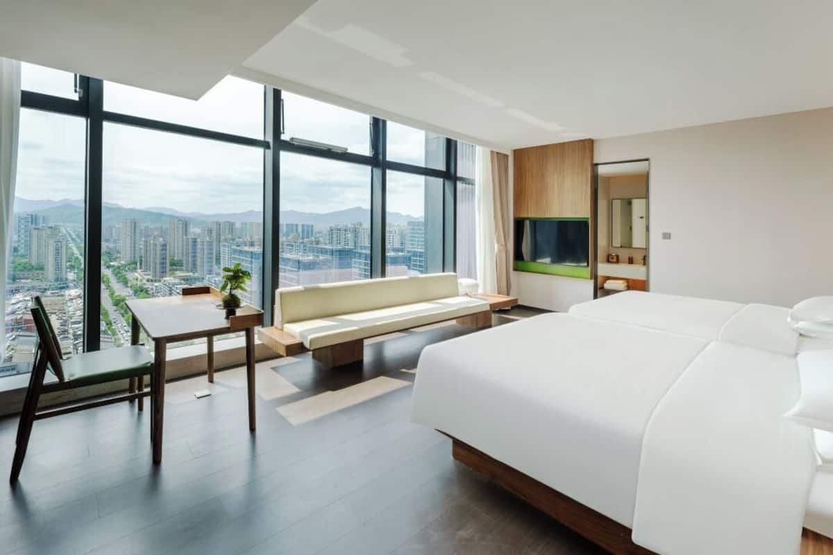 A modern hotel room with a large bed, a seating area with a table and chairs, and a large window offering a city view.
