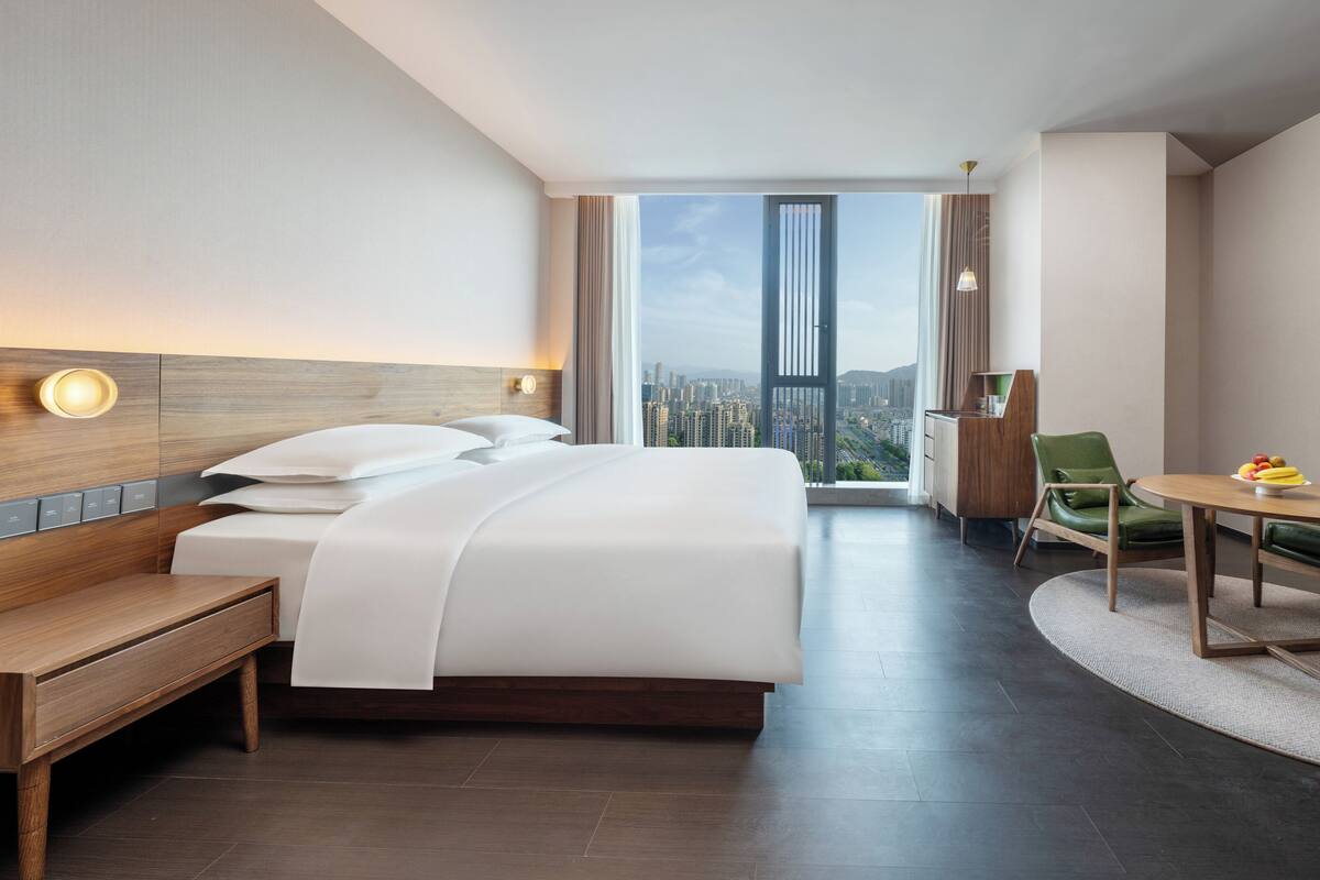 A modern hotel room with a large bed, a desk, a chair, and a view of a cityscape.