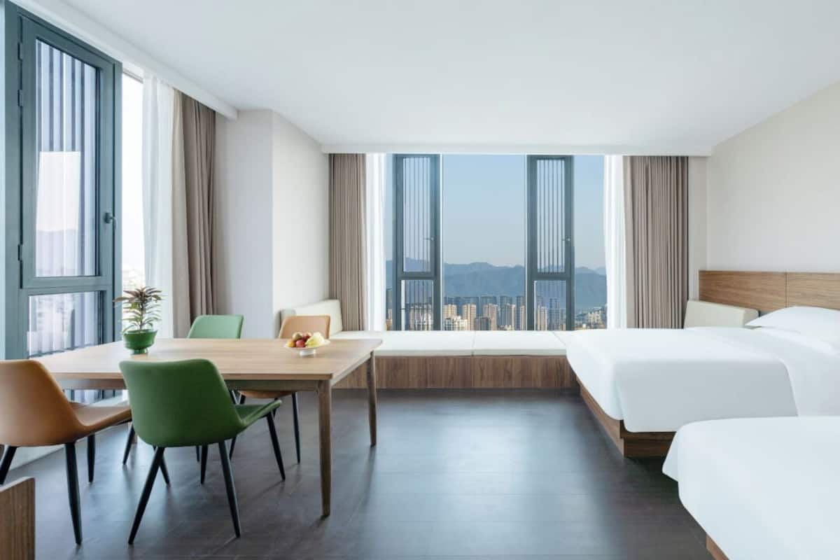 A hotel room with a large bed, a smaller bed, a desk with chairs, and a view of a cityscape through large windows.