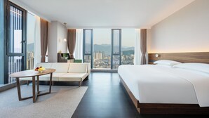 Habitación de hotel con una cama grande, un sofá, una mesita con fruta y vistas a un paisaje urbano a través de amplios ventanales.