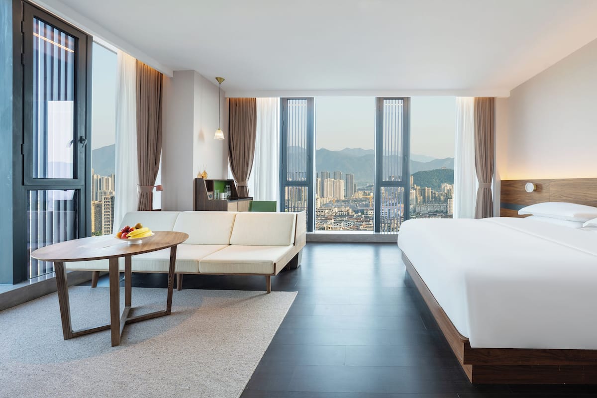 A hotel room with a large bed, a sofa, a small table with fruit, and a view of a cityscape through large windows.