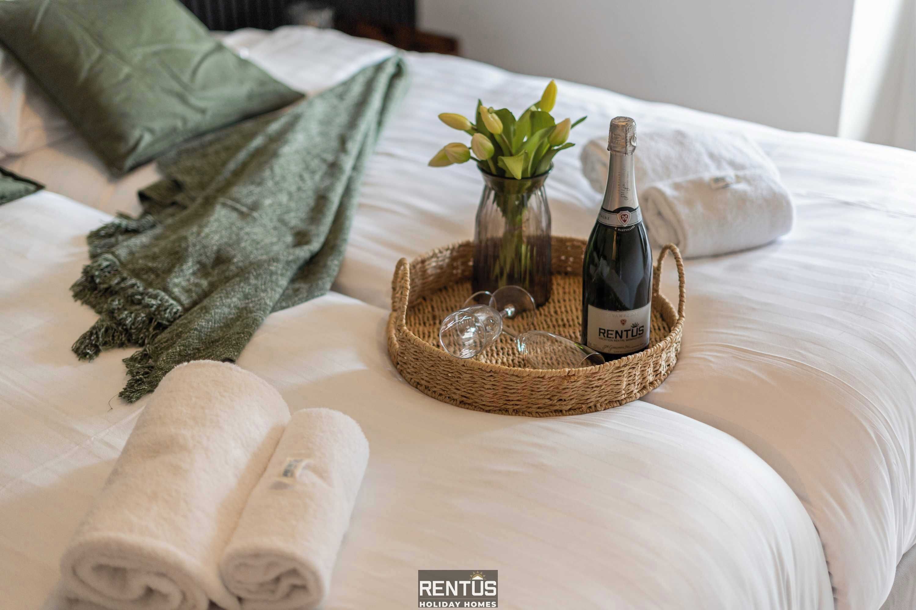 Una cama bien hecha con toallas blancas enrolladas, una botella de vino, una copa y un pequeño florero con hojas verdes.