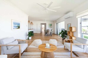 A spacious living area with a wooden floor, white sofas, a round wooden coffee table, and a ceiling fan. - The Dunes House - Oceanstays (Yamba)