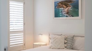 A bedroom with a bed, a nightstand with a lamp, a framed picture of a coastal landscape, and a window with blinds. - Eden - Oceanstays - Pet friendly (Yamba)