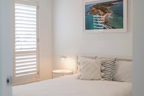 Une chambre à coucher avec un lit, une table de chevet avec une lampe, un tableau représentant un paysage côtier, et une fenêtre avec des stores.