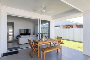 Un patio moderno con una mesa y sillas de madera, un ventilador de techo y un amplio ventanal que da a una sala de estar interior con sofá y televisor.