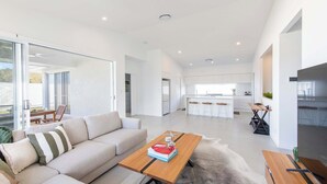 A modern living room with a sofa, a coffee table, and a dining area with a table and chairs. - Eden - Oceanstays - Pet friendly (Yamba)