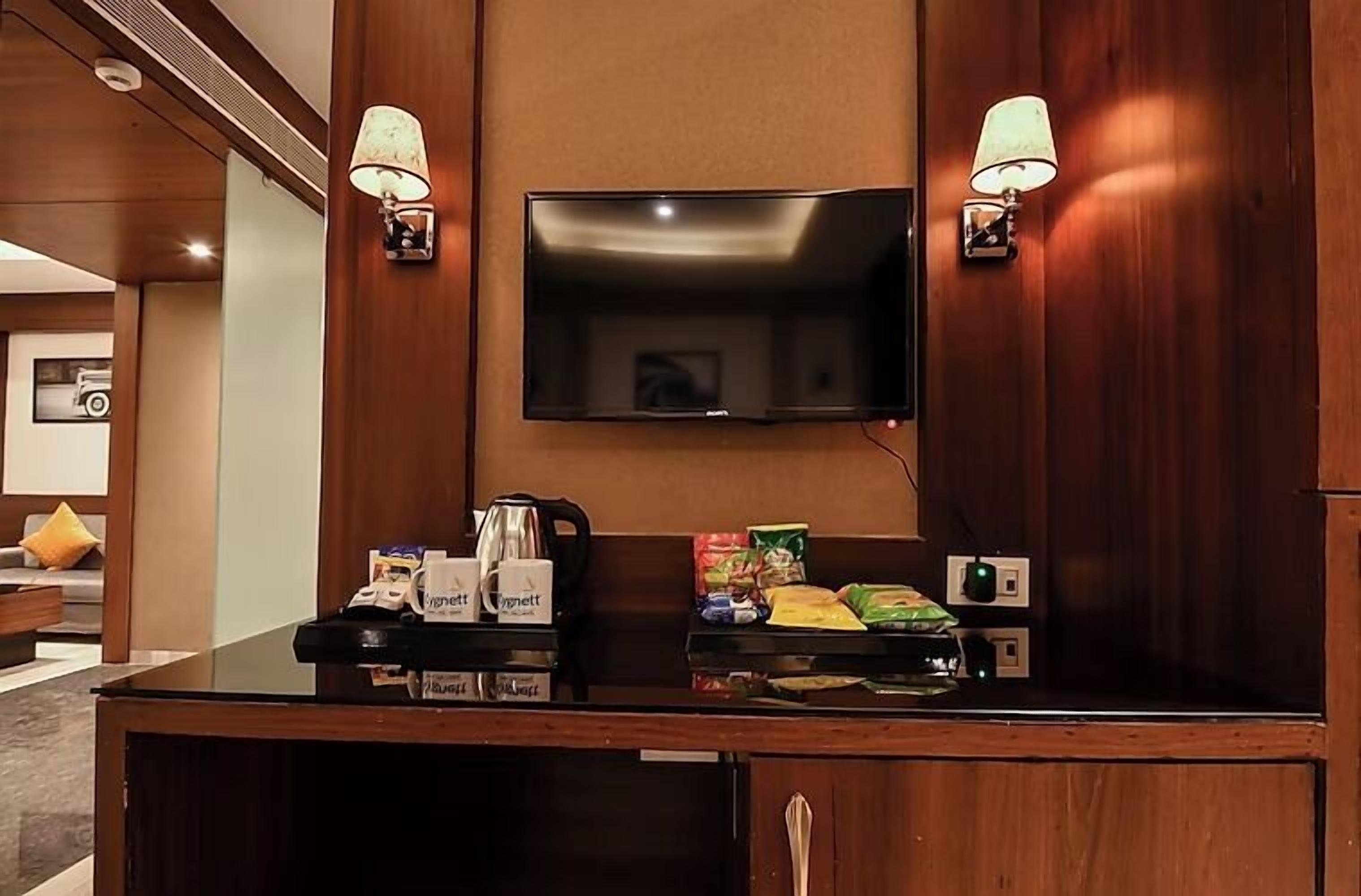 A hotel reception area with a flat-screen TV mounted on the wall, two wall-mounted lamps, a coffee station with branded cups, and a snack tray.
