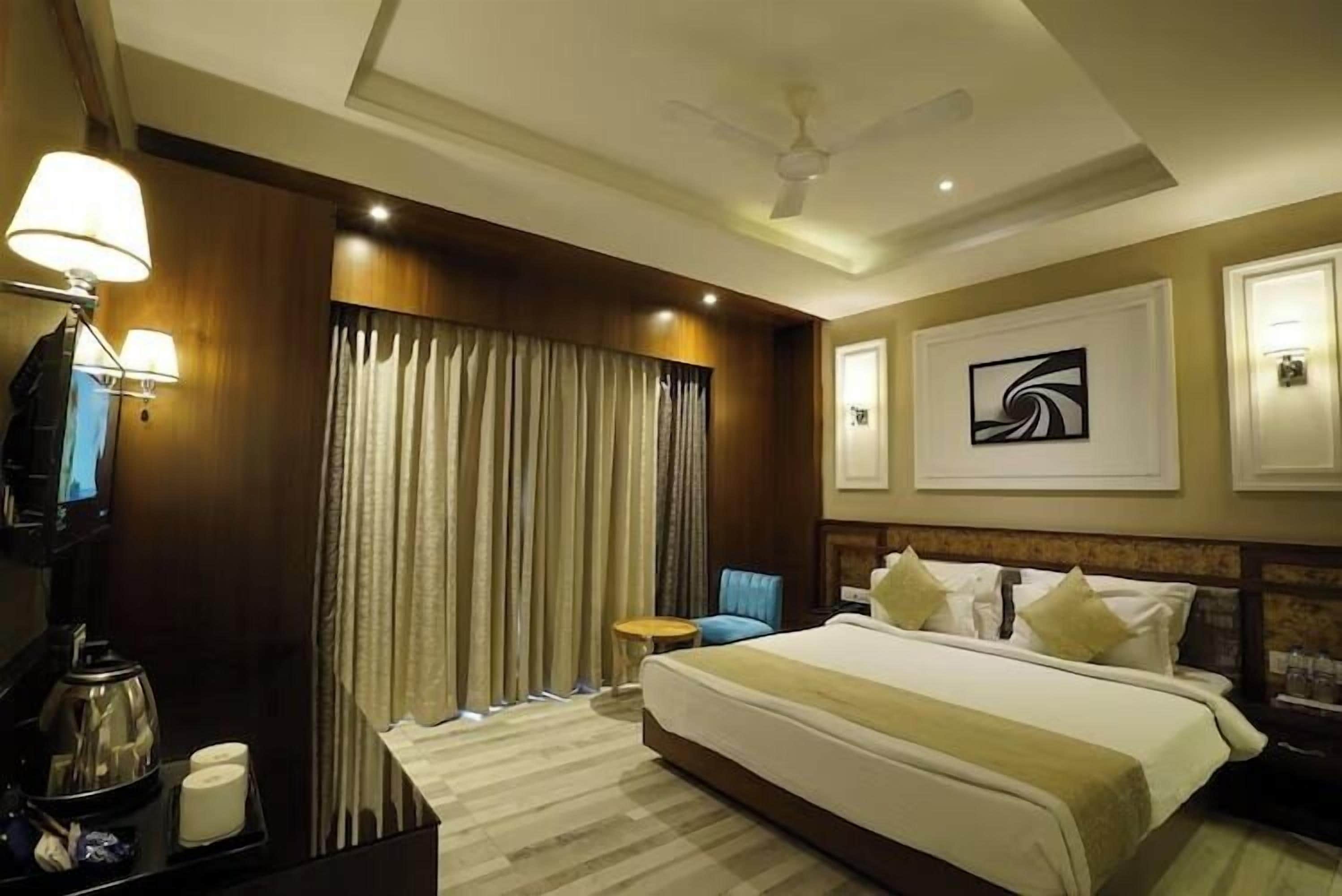 Une chambre d’hôtel avec un grand lit, un bureau avec une lampe, une chaise, un ventilateur de plafond et un tableau au mur.