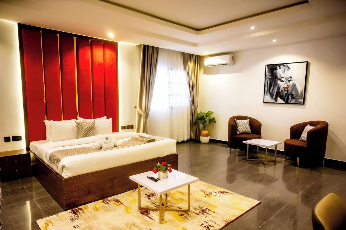 A modern hotel room with a large bed, a small table, two armchairs, a potted plant, and a painting on the wall.