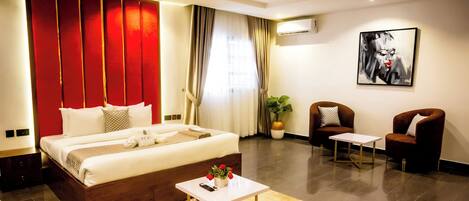A modern hotel room with a large bed, a small table, two armchairs, a potted plant, and a painting on the wall.
