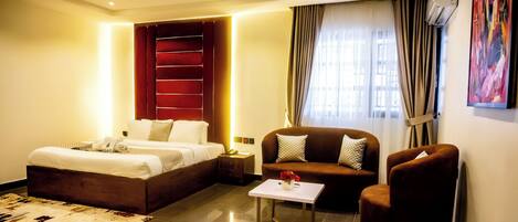 A hotel room with a large bed, a sofa, a small table, and a chair. There is a window with curtains and an air conditioning unit on the wall.