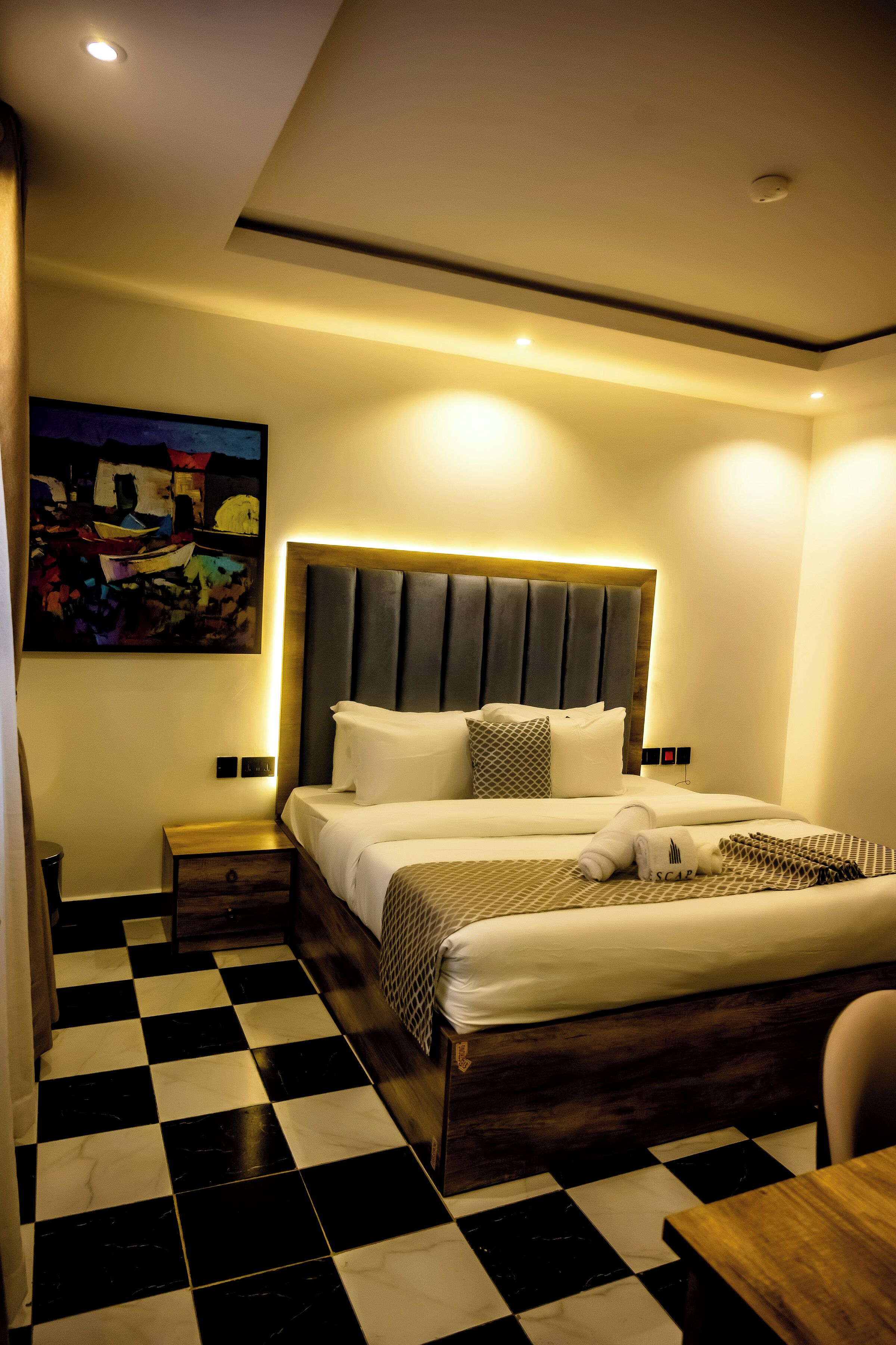 Una habitación de hotel moderna con piso de cuadros, una cama grande con cabecera de madera, una mesita de noche y un cuadro colgado en la pared.