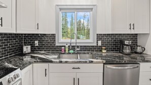 A modern kitchen with white cabinets, a black and white marble countertop, a stainless steel dishwasher, and a window with a view of trees. - Comfortable Cottage with hot tub in Delta Junction, AK (Delta Junction)
