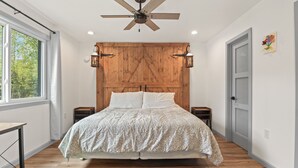 A bedroom with a wooden headboard, a ceiling fan, two bedside tables, and a large window. - Comfortable Cottage with hot tub in Delta Junction, AK (Delta Junction)