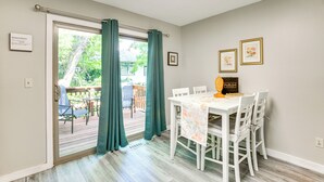 Un área de comedor con una mesa y sillas blancas, una ventana con cortinas verdes y vista a una terraza al aire libre.
