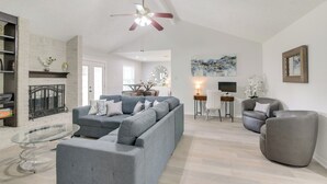 A spacious living room with a grey sectional sofa, a glass coffee table, and a fireplace. There is a dining area with a table and chairs, and a TV mounted on the wall. - 30 Miles to NRG Stadium - Family Home in Houston! (Houston)