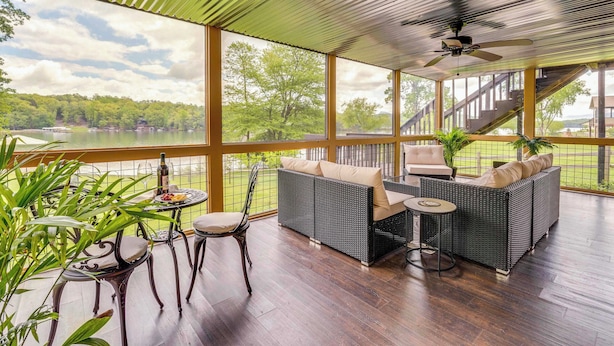 A covered outdoor seating area with wicker furniture, a table, and chairs, overlooking a lake and greenery. - Lake Chatuge Home: Boat Dock, Hot Tub & Perks! (Hayesville)