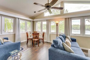 A spacious room with a large window, a ceiling fan, a dining area with chairs and a table, and a blue sofa with cushions. - Lake Chatuge Home: Boat Dock, Hot Tub & Perks! (Hayesville)
