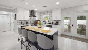 A modern kitchen with white cabinets, a central island with bar stools, and a view of the outdoors through glass doors. - Coastin Close Family Fun HTD Pool Outdoor Games (Pinellas Park)