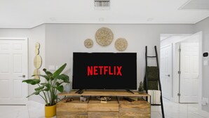 A modern living room with a large flat-screen TV displaying the Netflix logo, a wooden entertainment unit, a potted plant, and a ladder against the wall. - Coastin Close Family Fun HTD Pool Outdoor Games (Pinellas Park)
