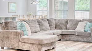 A large L-shaped sectional sofa with multiple cushions, a dining table with chairs, and large windows with blinds.