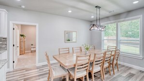 A dining area with a wooden table and chairs, a hanging light fixture, and a large window with blinds.