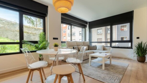 A modern living room with a round table, chairs, a sofa, and a coffee table. Large windows provide views of the outside. - Diseño y confort en Via Cornelia (Santander)