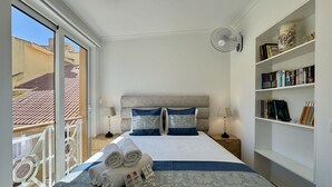 Habitación de hotel con cama, dos mesitas de noche, una estantería y un balcón con barandilla.