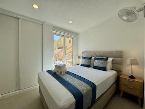 Habitación de hotel con cama, mesita de noche, lámpara y ventana con vistas a edificios.