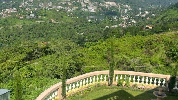 A lush green landscape with a mountainous backdrop and a view of a town below. - The Taal Cottage (Bhimtal)