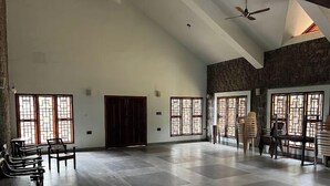 A spacious room with a high ceiling, large windows with intricate designs, and a stone wall. There are several wooden chairs and tables arranged for seating. - Joppa House (Nagercoil)
