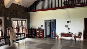 A spacious room with a high ceiling, stone walls, and a wooden staircase. There are several wooden chairs and a small table, along with a cabinet and a door leading to another room. - Joppa House (Nagercoil)