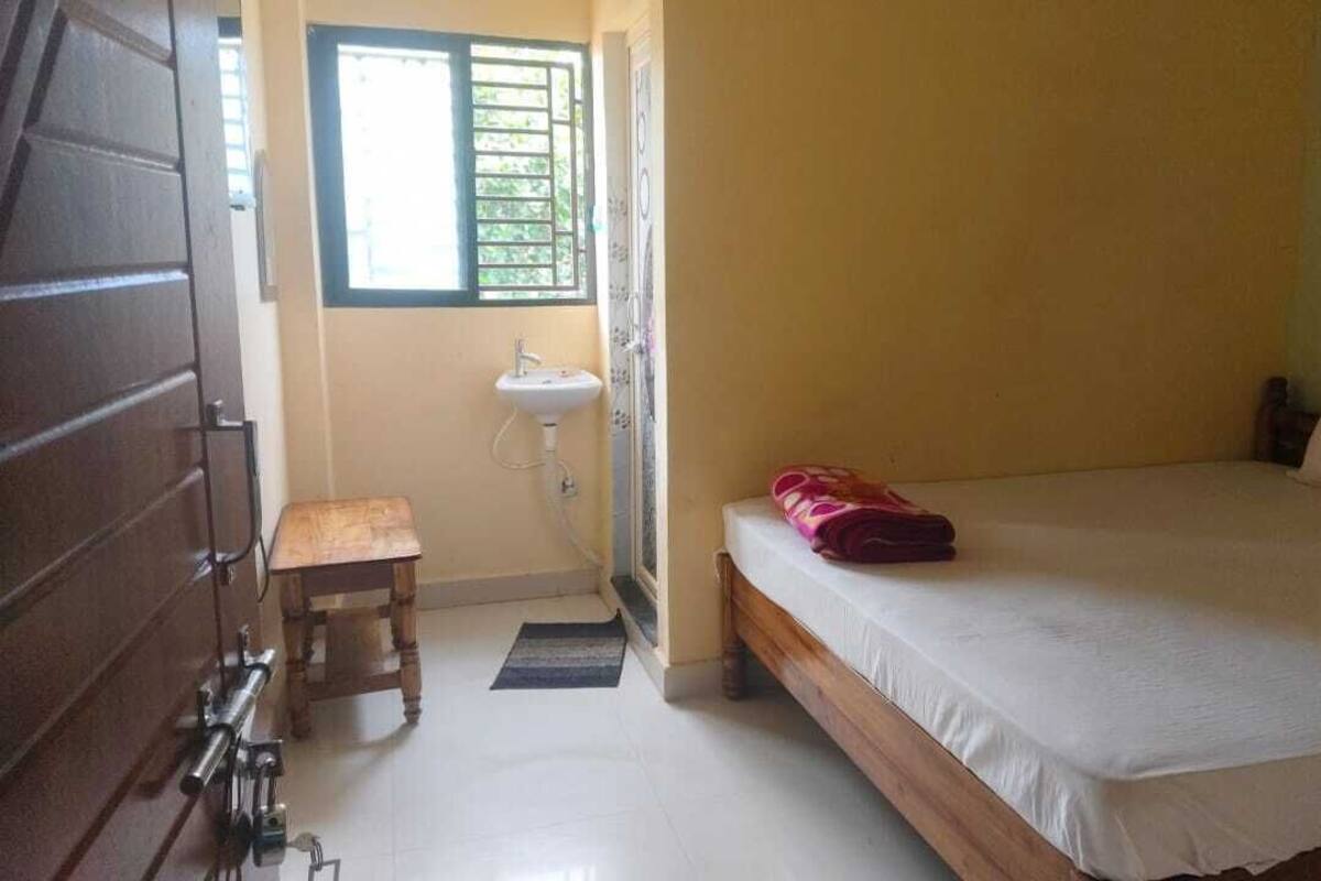 A room with a bed, a small wooden stool, a sink, and a window with bars.