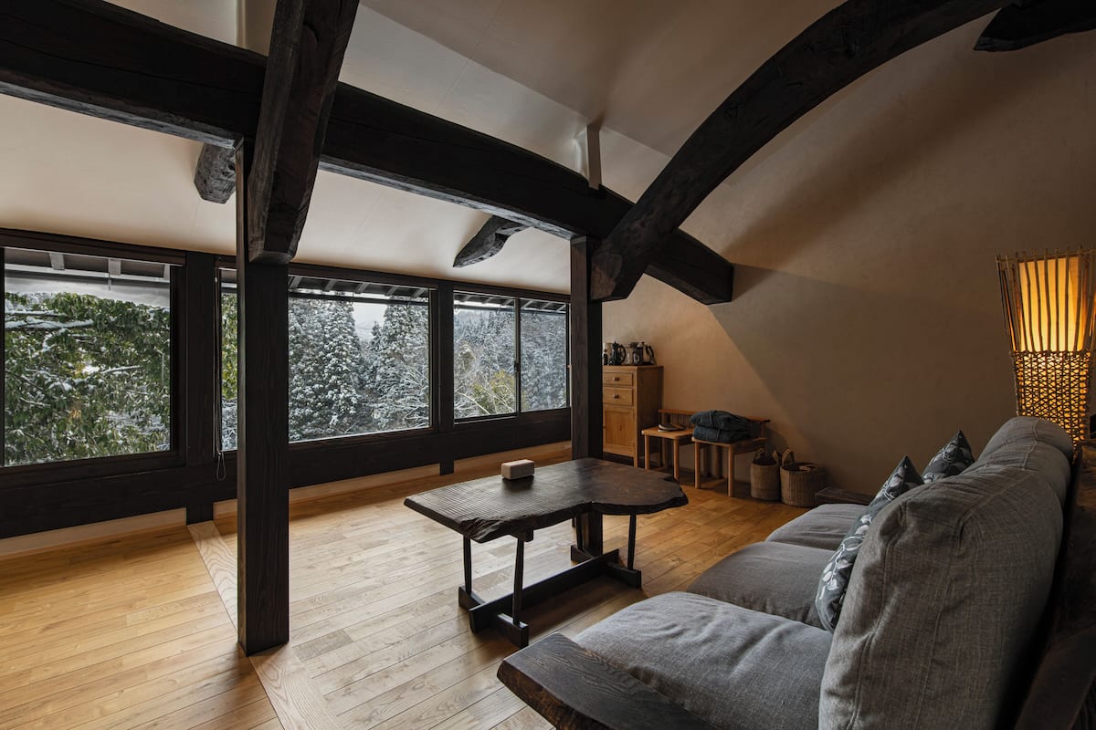 Een kamer met houten vloeren, een bank, een tafel en grote ramen met uitzicht op bomen.