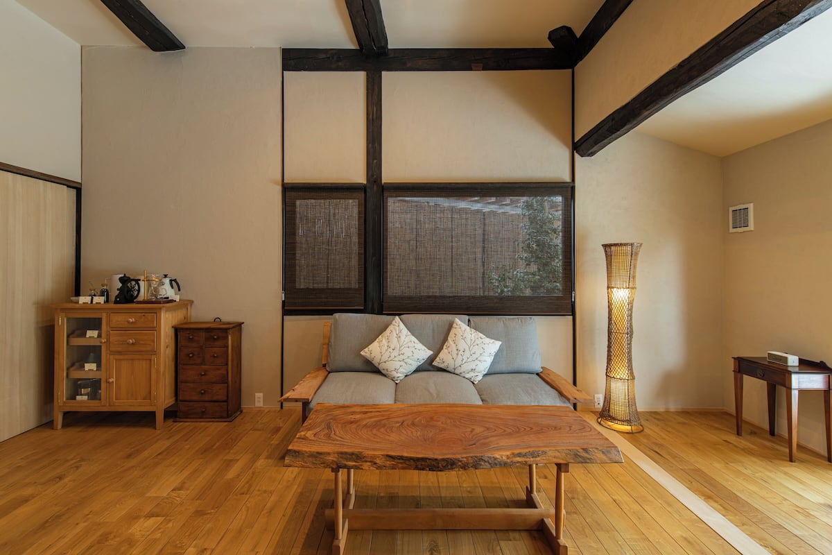 Een kamer met een houten vloer, een bank met kussens, een houten tafel, een kast en een hoge staande lamp.