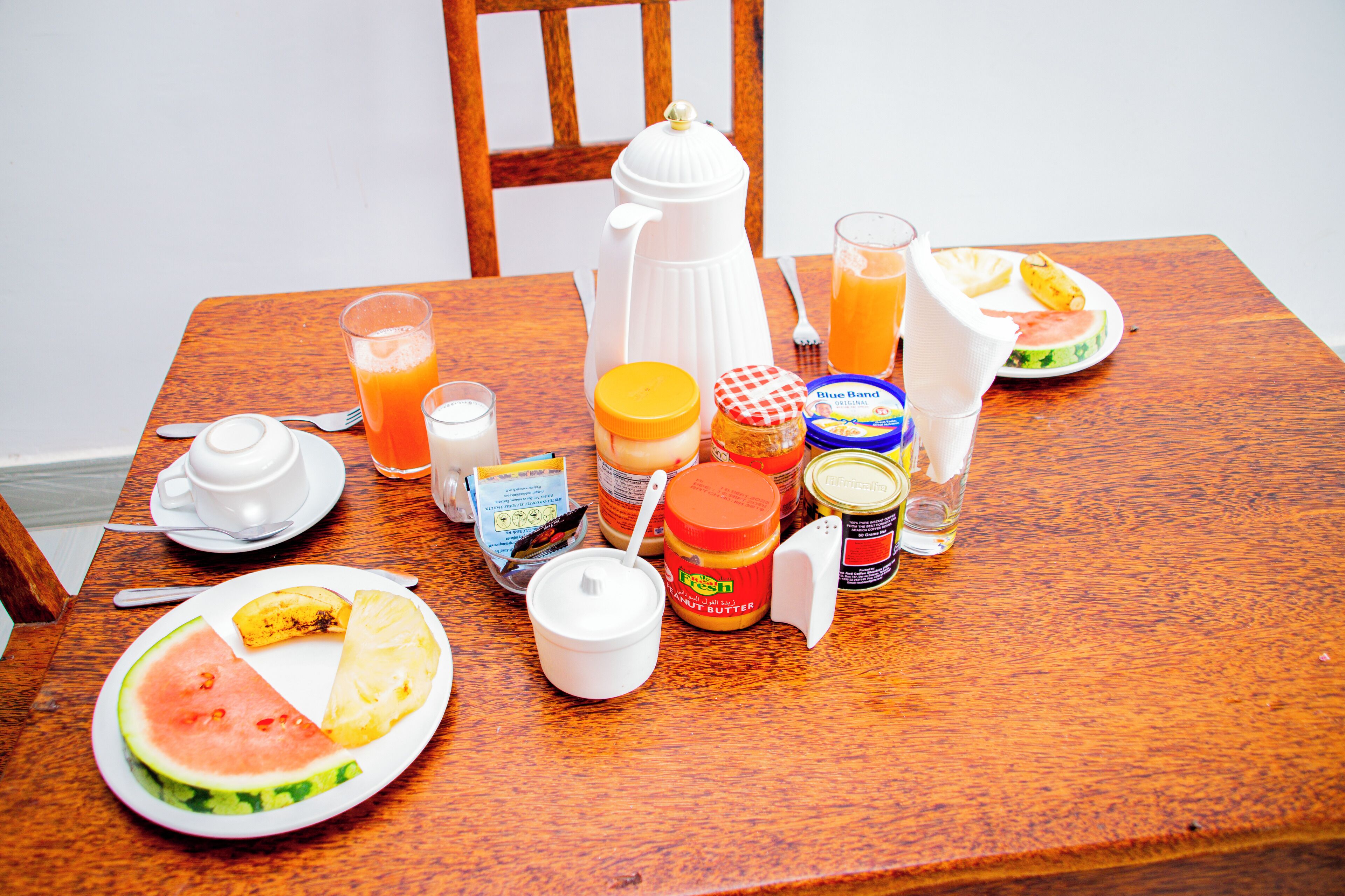 Et træbord med morgenmadsretter, herunder en tallerken med vandmelon og ananas, en kaffepotte, et glas appelsinjuice og diverse tilbehør.