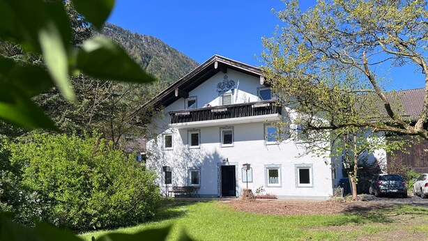 Een gebouw met twee verdiepingen, met een balkon, omgeven door groen en een berg op de achtergrond.