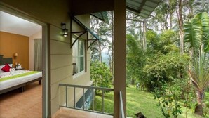 A room with a bed, a small table with a lamp, and a view of a lush garden.